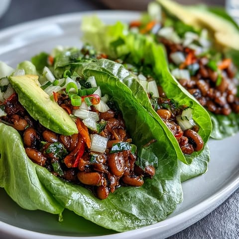 A close-up of a hand holding a crisp Black-Eyed Pea Lettuce Wrap, showcasing vibrant cilantro and a drizzle of hot sauce.